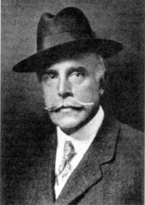 Black and white headshot of an older man in a suit, with a mustache and fedora hat