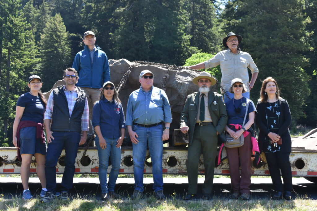 a group of people pose standing in front of a truck bed where a huge rock is strapped down