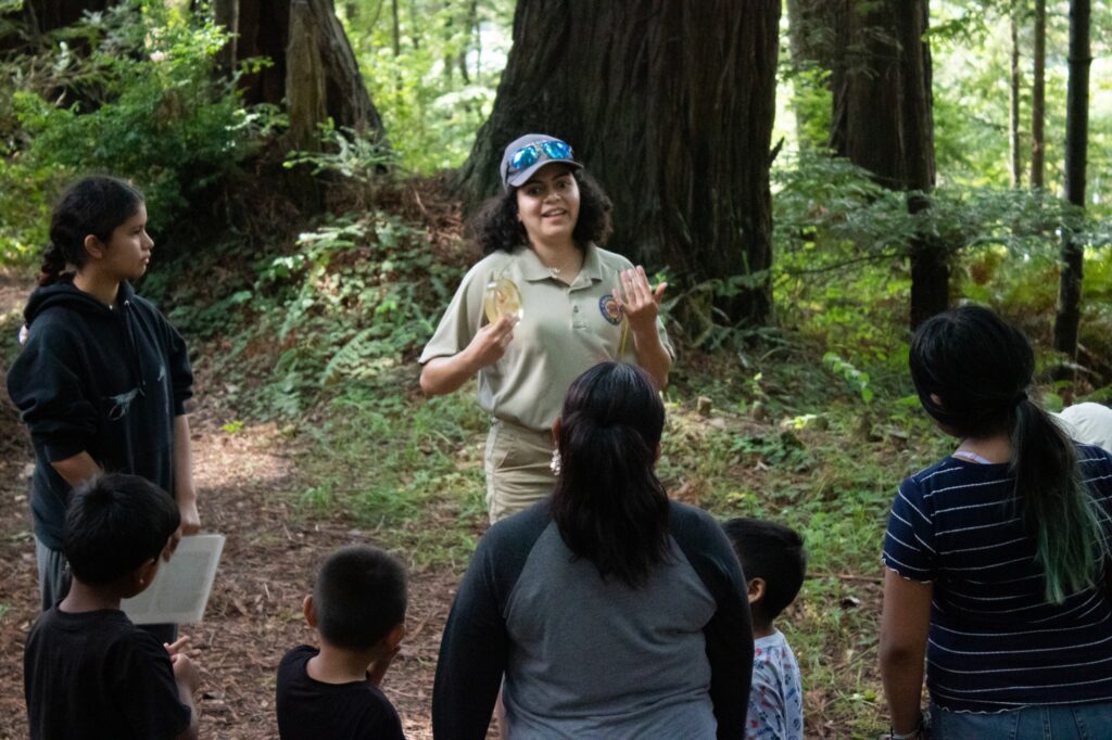 a woman with a cap is explaining to a small crowd of people while standing in front of a large tree trunk
