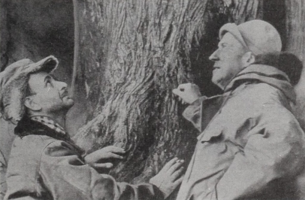 black and white medium shot of two men, shoulder up, touching the bark of a tree and looking in different directions