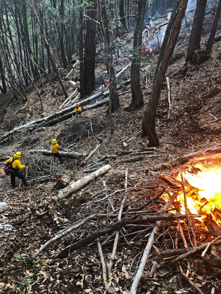 a slope that goes downward toward the right, where many workers wearing yellow vests are hard hat are positioned as a fire burns on the left side of the photograph