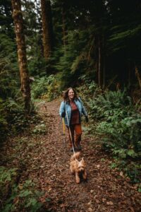 a person walks with a walking stick and a dog leashed to them through a forested trail