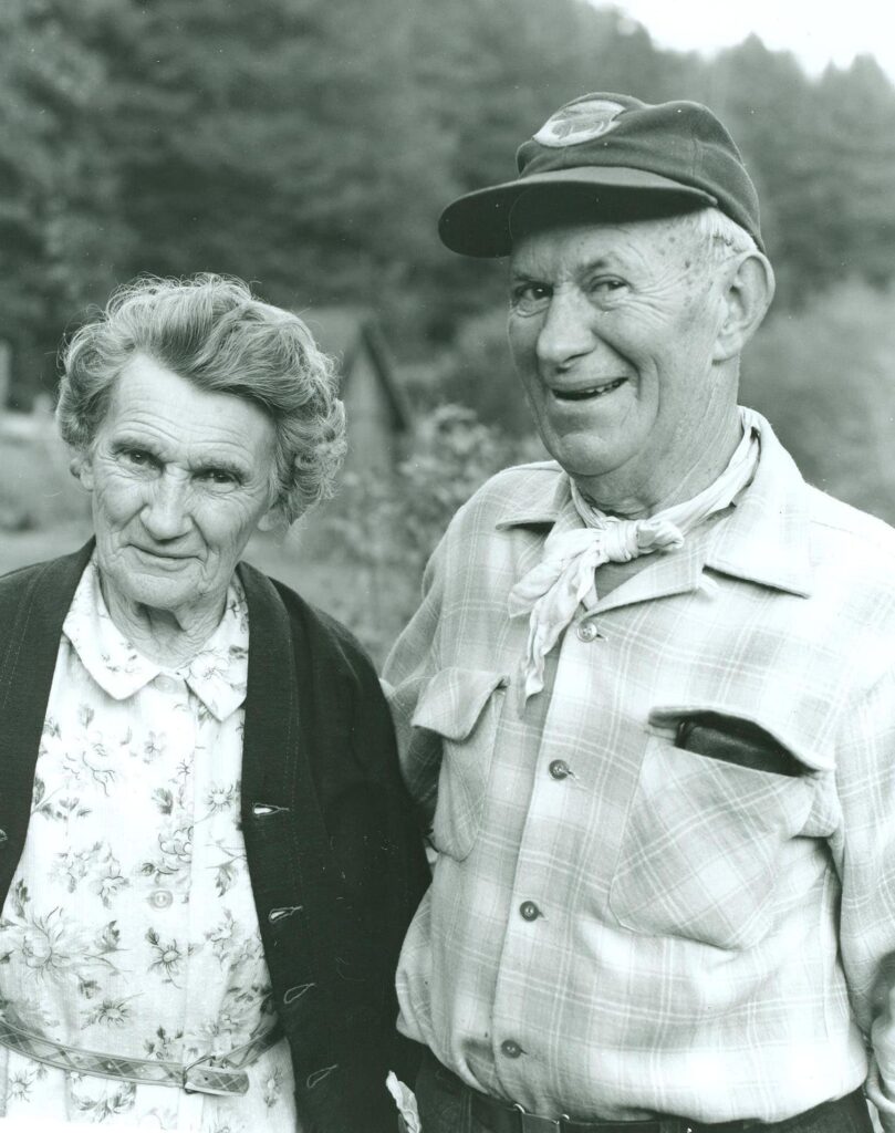 An older woman in a flowered dress stands next to an older man in a plaid shirt and neckerchief.