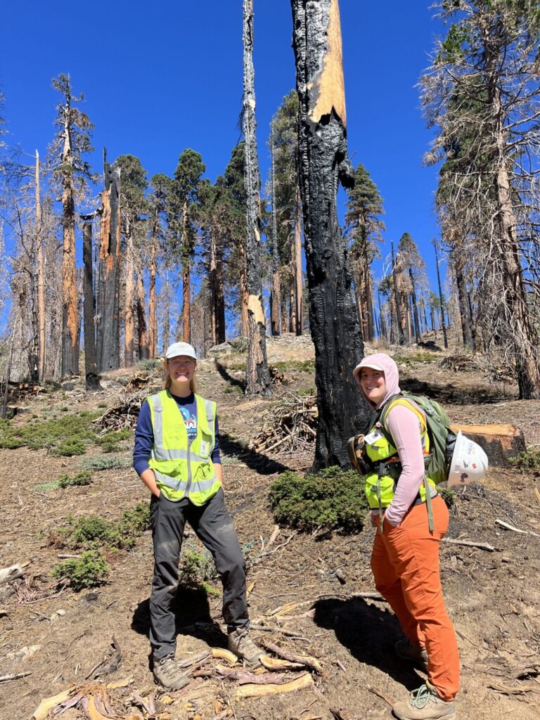 Two female restoration workers in neon-green vests smile in front of a partially burned sequia forest