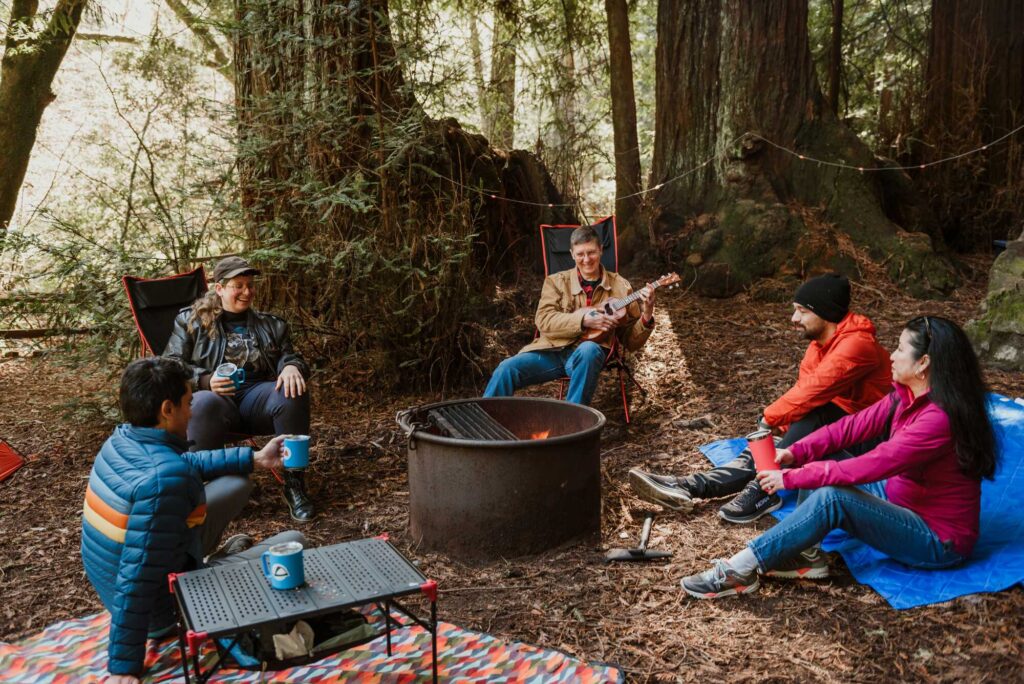 A person playing a guitar sits at a campfire ring with four other campers sitting beneath large redwood trees