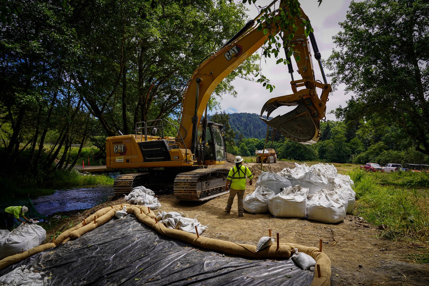 A large excavator on a river bank stands next to a pile of white bags while a person in hi-viz uniform looks on