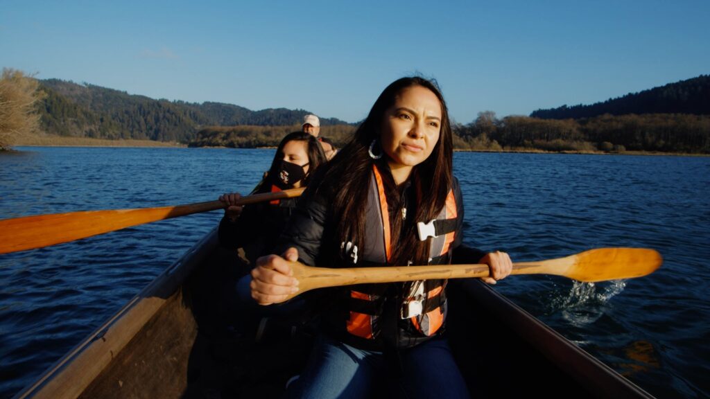 Three people paddle a canoe on a large river, with woman holding a wooden paddle in the foreground