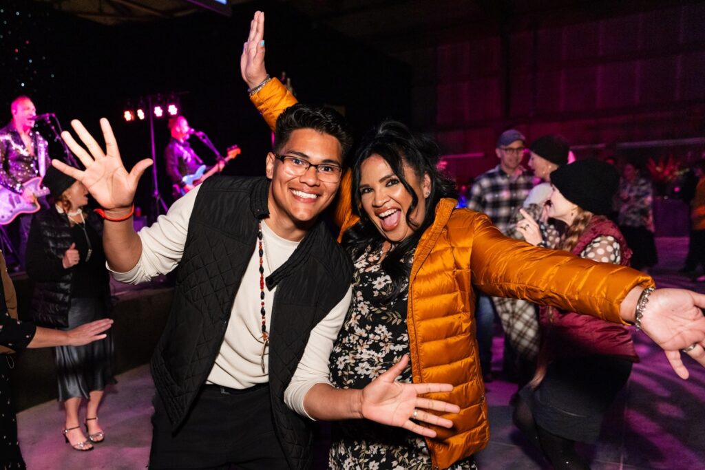 Two people in puffy jackets smile broadly at the camera from the dance floor with their hands outstretched
