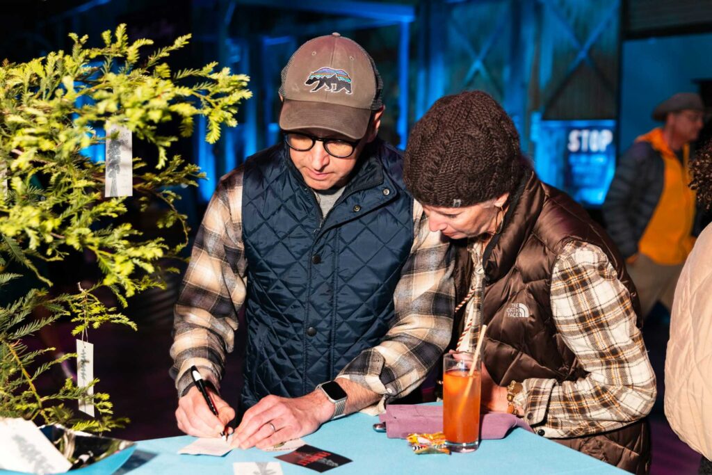 Two people wearing flannel write on pieces of paper next to a potted tree with tags dangling off its branches