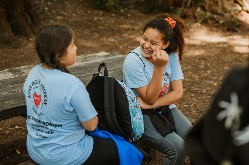 Two girls wearing Asisa Pacific American community Center T-shirts laugh while sitting on a bench in the forest
