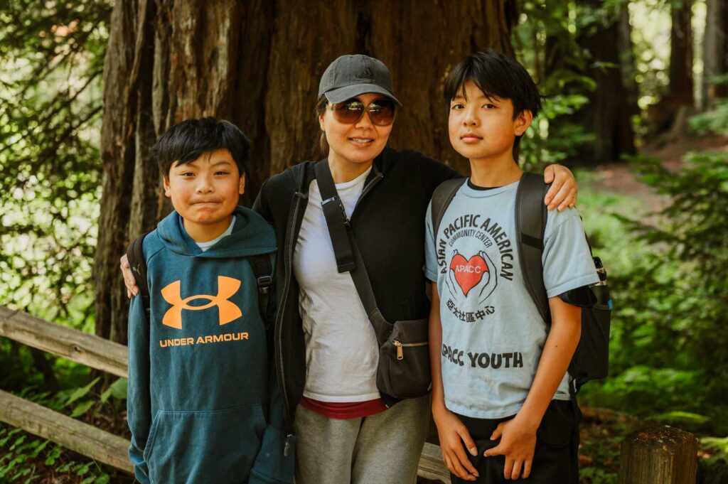 An Asian woman wearing sunglasses has her arms around the shoulders of two young boys, one of whom wears an Asian Pacific American Community Center T-shirt.