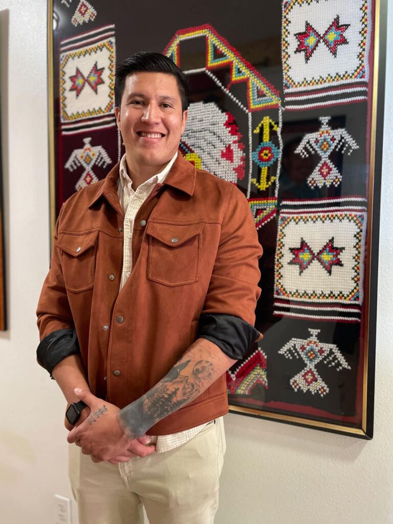 A man wearing a white shirt and rust brown jacket poses with sleeves rolled up to reveal a quarter sleeve tattoo on his forearm