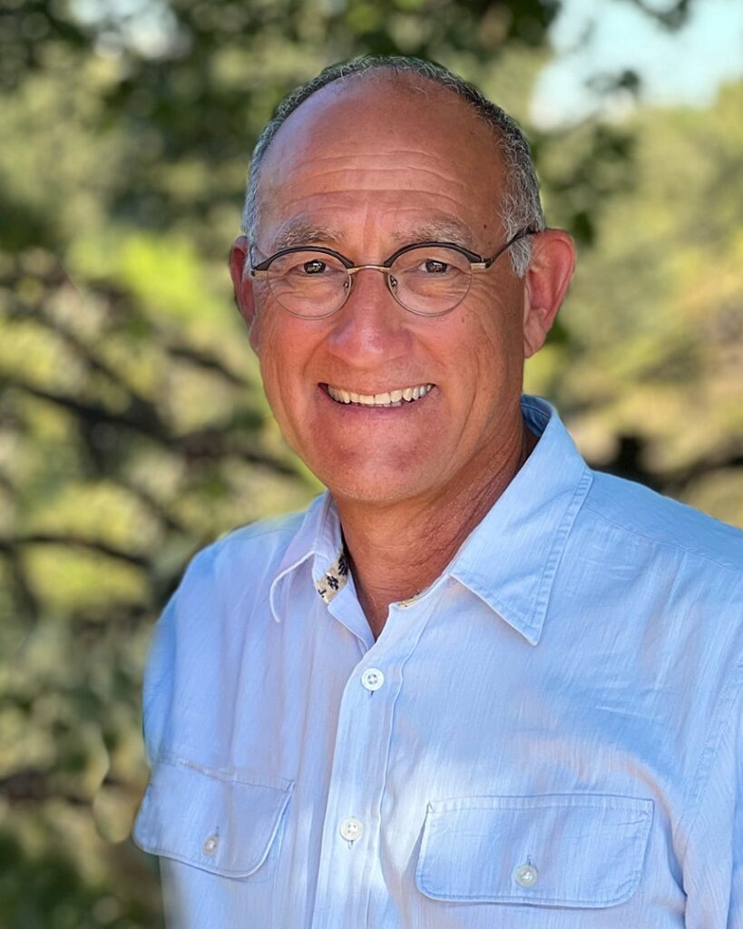 An older man wearing round-rimmed glasses smiles at the camera while wearing a blue shirt.
