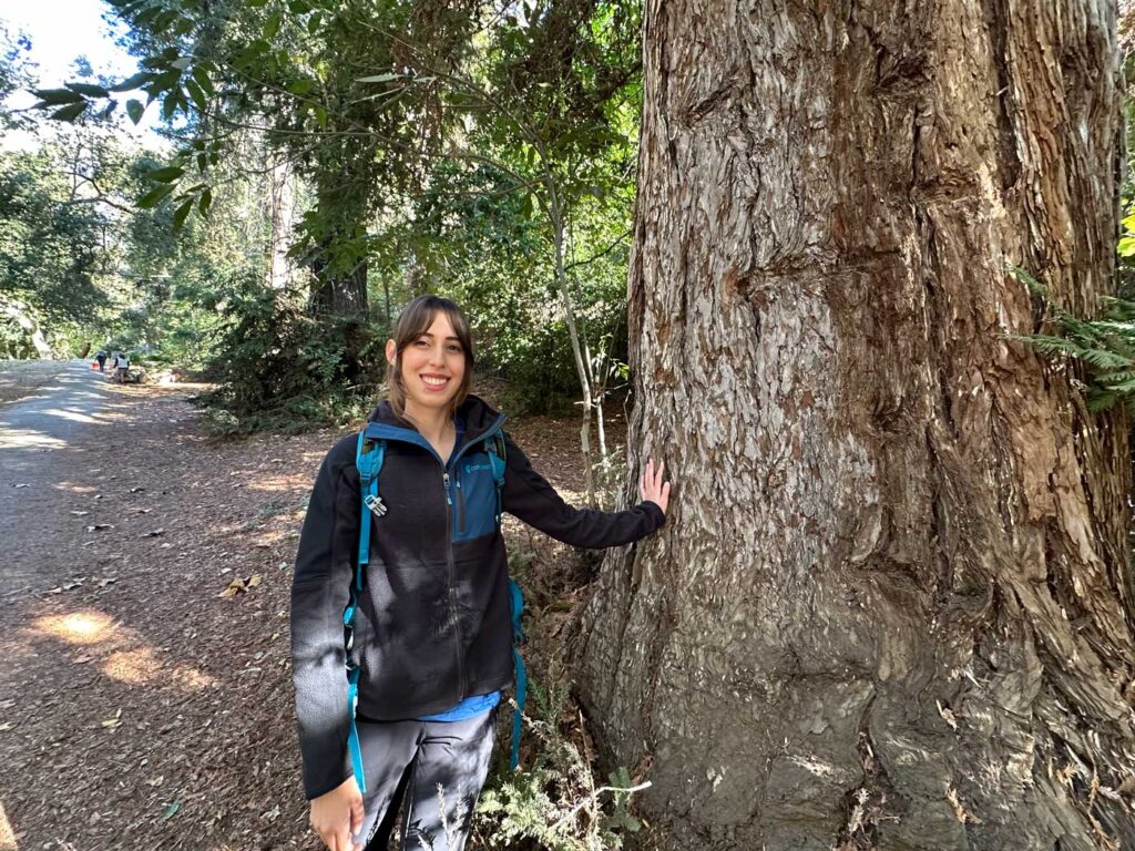 A woman wearing a backpack smiling with her hand on the trunk of a redwood tree.