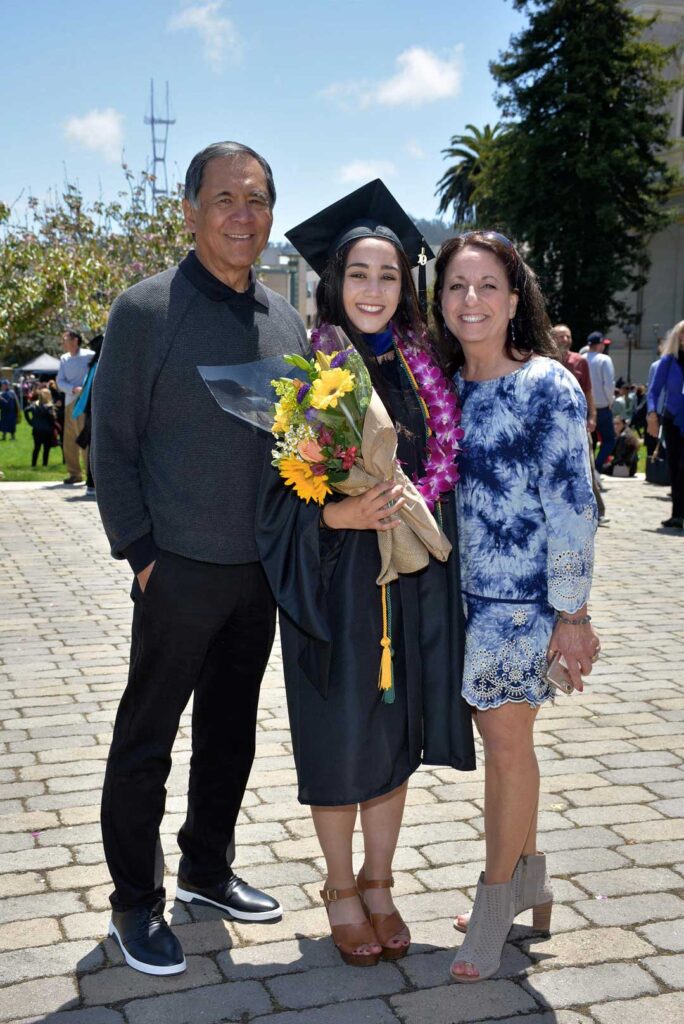 A young woman in a graduation gown and cap holding a bouquet and standing between her mother and father.