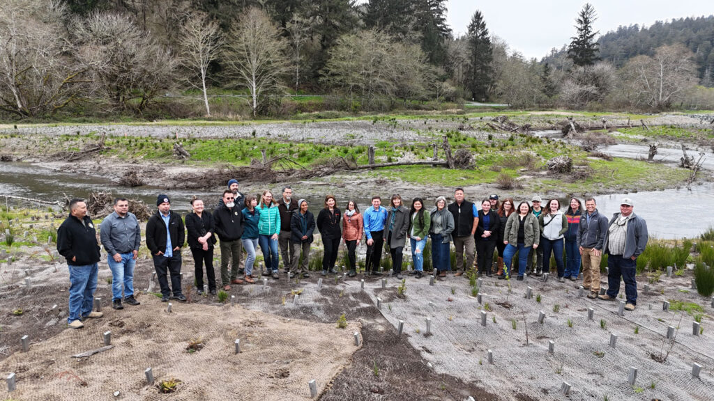 26 people standing outside in floodplains where small wood pieces are marking the ground