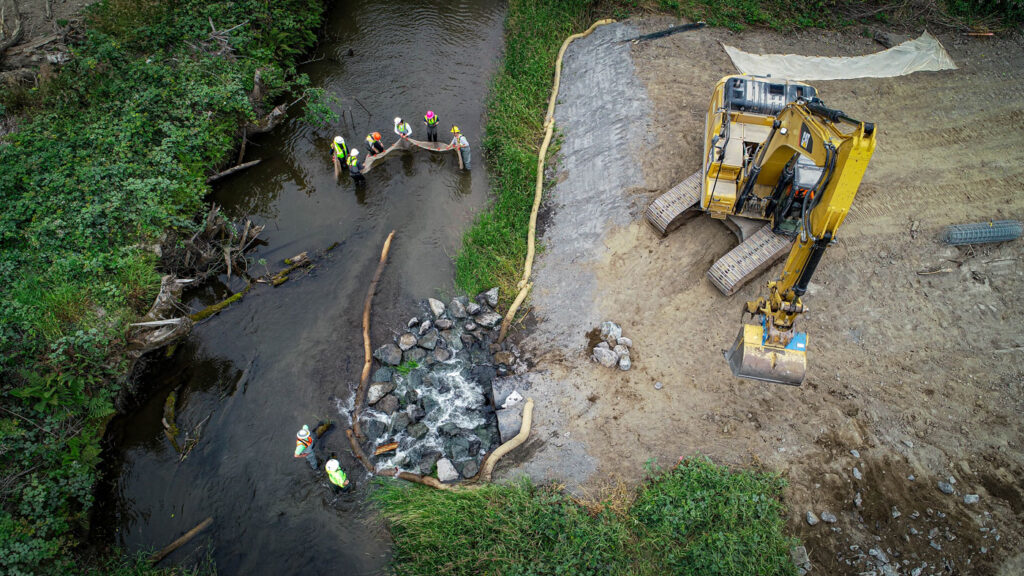 Overhead aerial view of a river bank, with an excavator on the sand as people are standing in the water and wearing hi-viz clothing