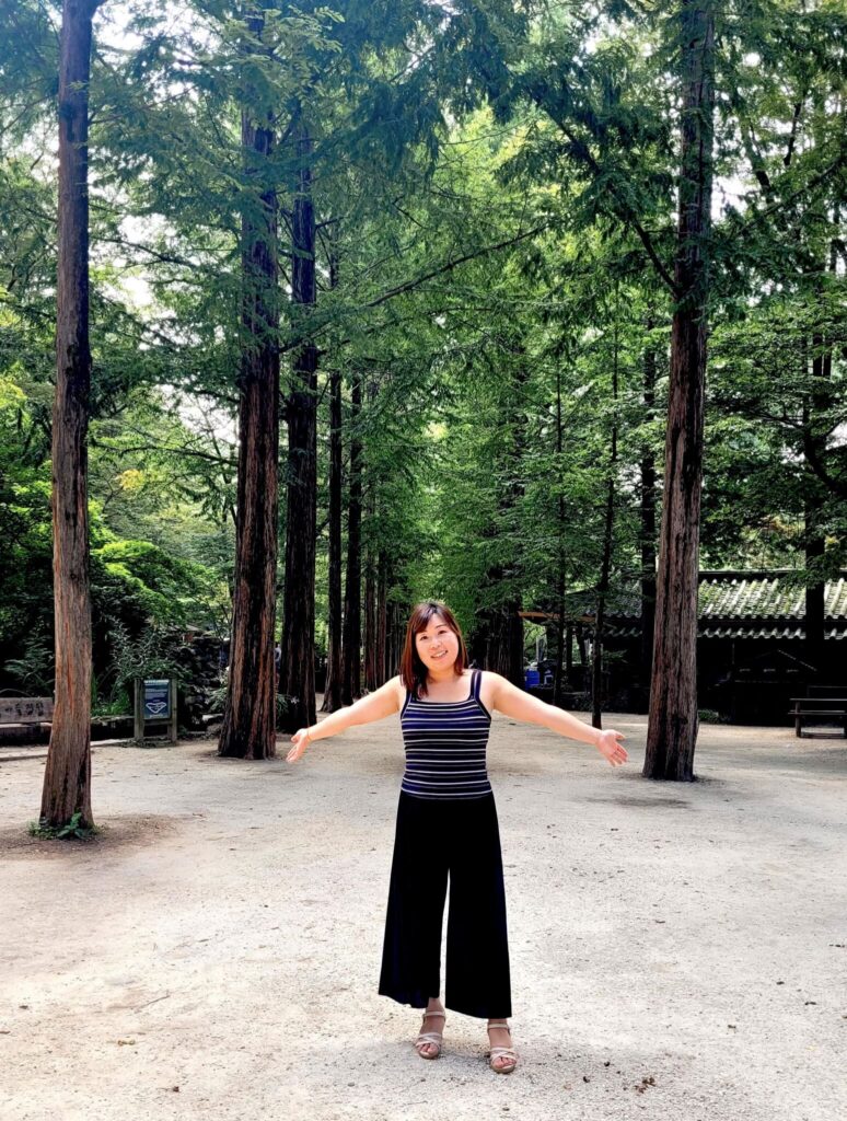A smiling young Asian woman stands with her arms outstretched at the entrance to a lane lined with tall trees.