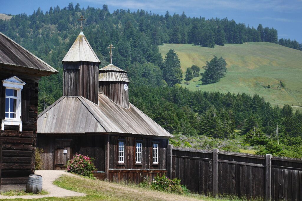 Photograph of a Russian Orthodox church, with a wooden fence and green hills in the background.