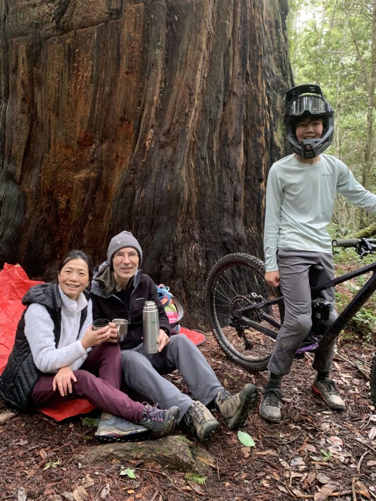 A teenage boy wearing a mountain biking helmet and straddling a mountain bike, standing in front of a giant sequoia. His parents are seated nearby in camping gear, holding a thermos and cup.