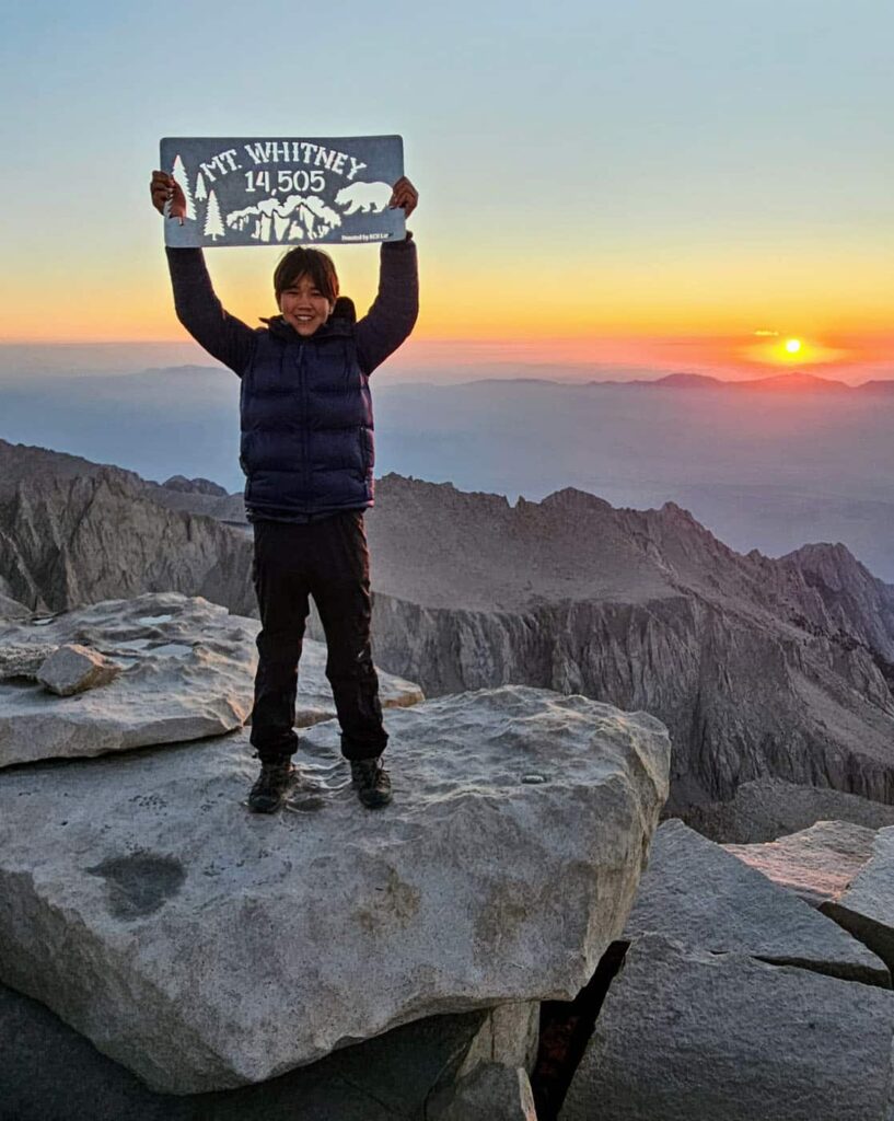 A teenage boy wearing a down jacket standing at the summit of Mt. Whitney at sunset, holding a sign that says Mt. Whitney, 14,505'.