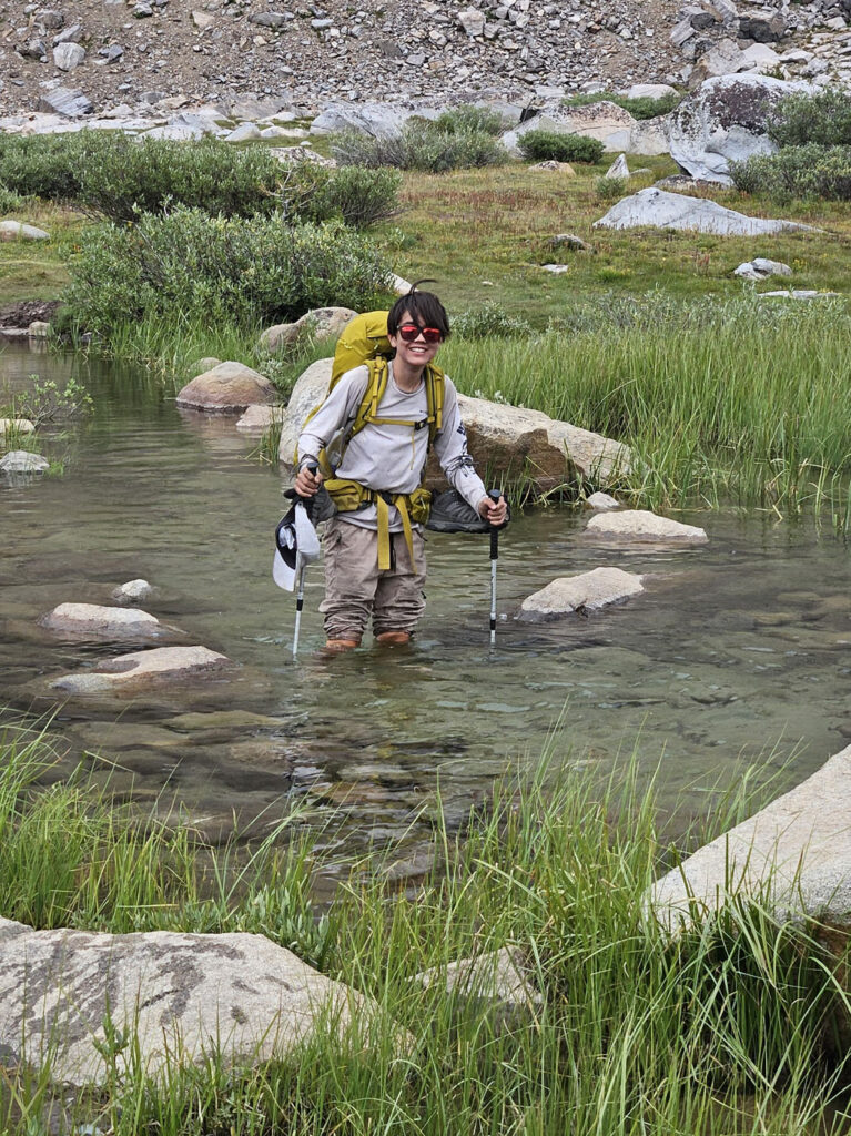A teenaged boy wearing a backpackers hiking rucksack and sunglasses, holding trekking poles, and is standing knee deep in a stream filled with small boulders and tall grasses.