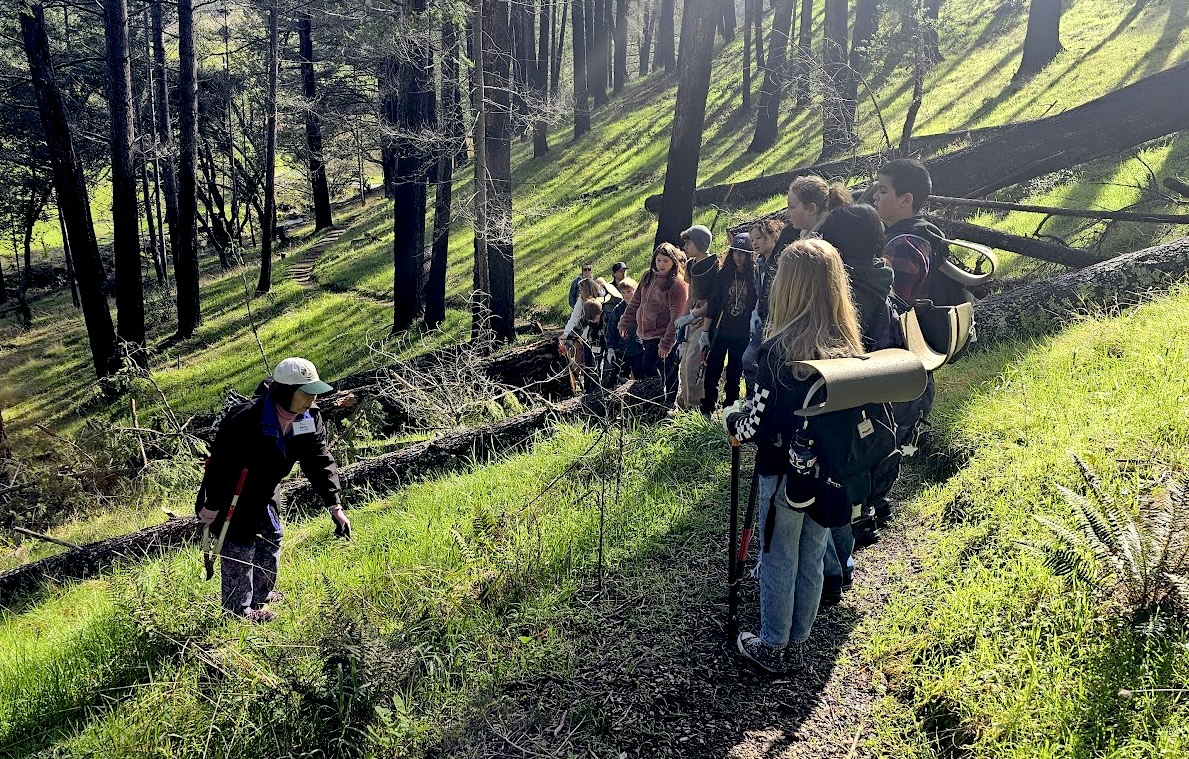 Kids on a hillside path listen to a man wearing a cap who stands to the left on top of grass