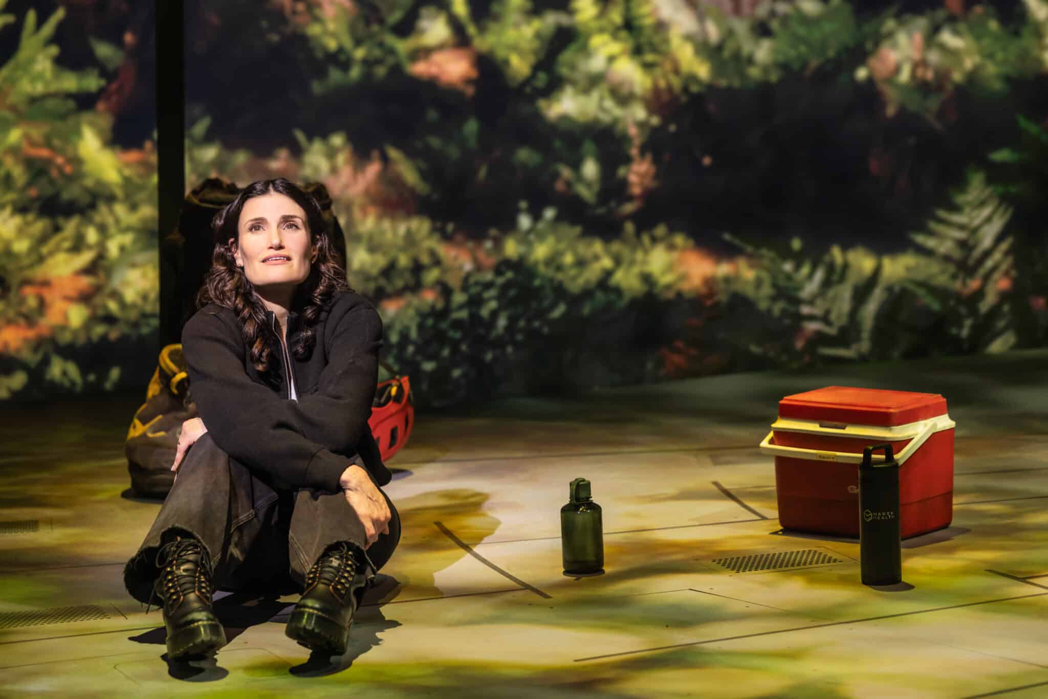 A woman sits on a stage decorated as a forested area, with a thermos and cooler by her side, gazing into the distance.