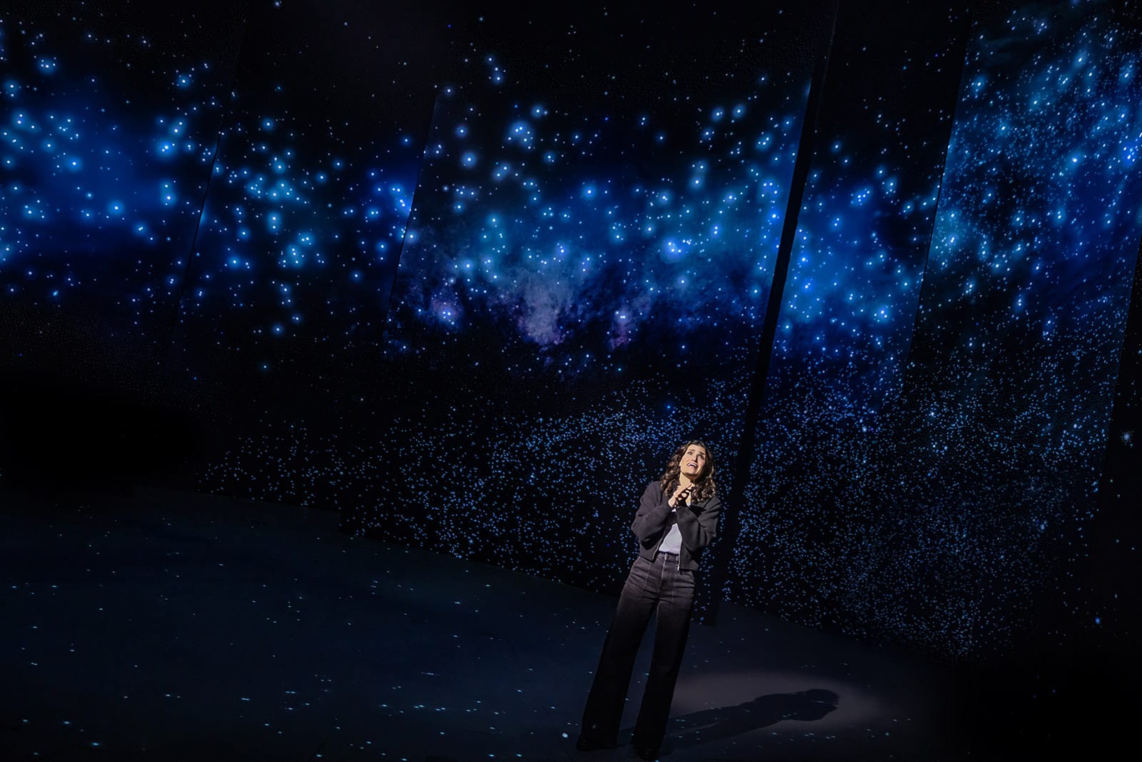 A woman stands on a dark stage beneath a sweeping, star-filled backdrop, looking upward with her hands clasped near her chest. She’s dressed in black pants and a jacket, and the blue glow of the projected stars surrounds her.