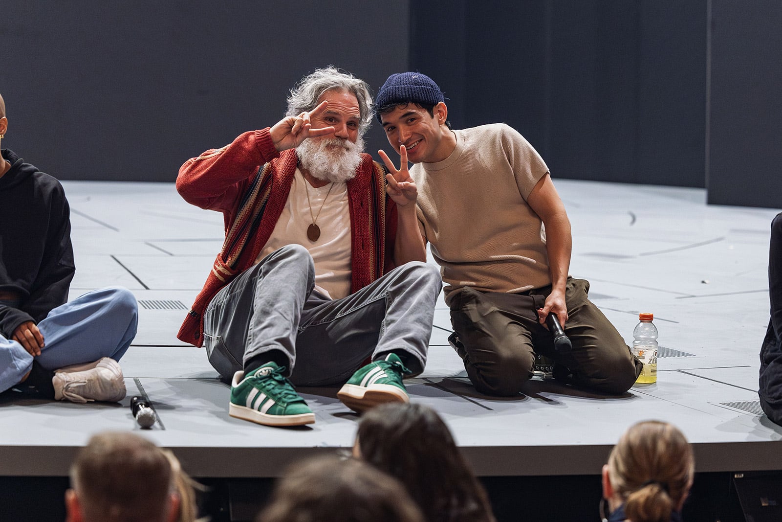 A man and an older man sit on a stage, posing with their fingers in a V shape