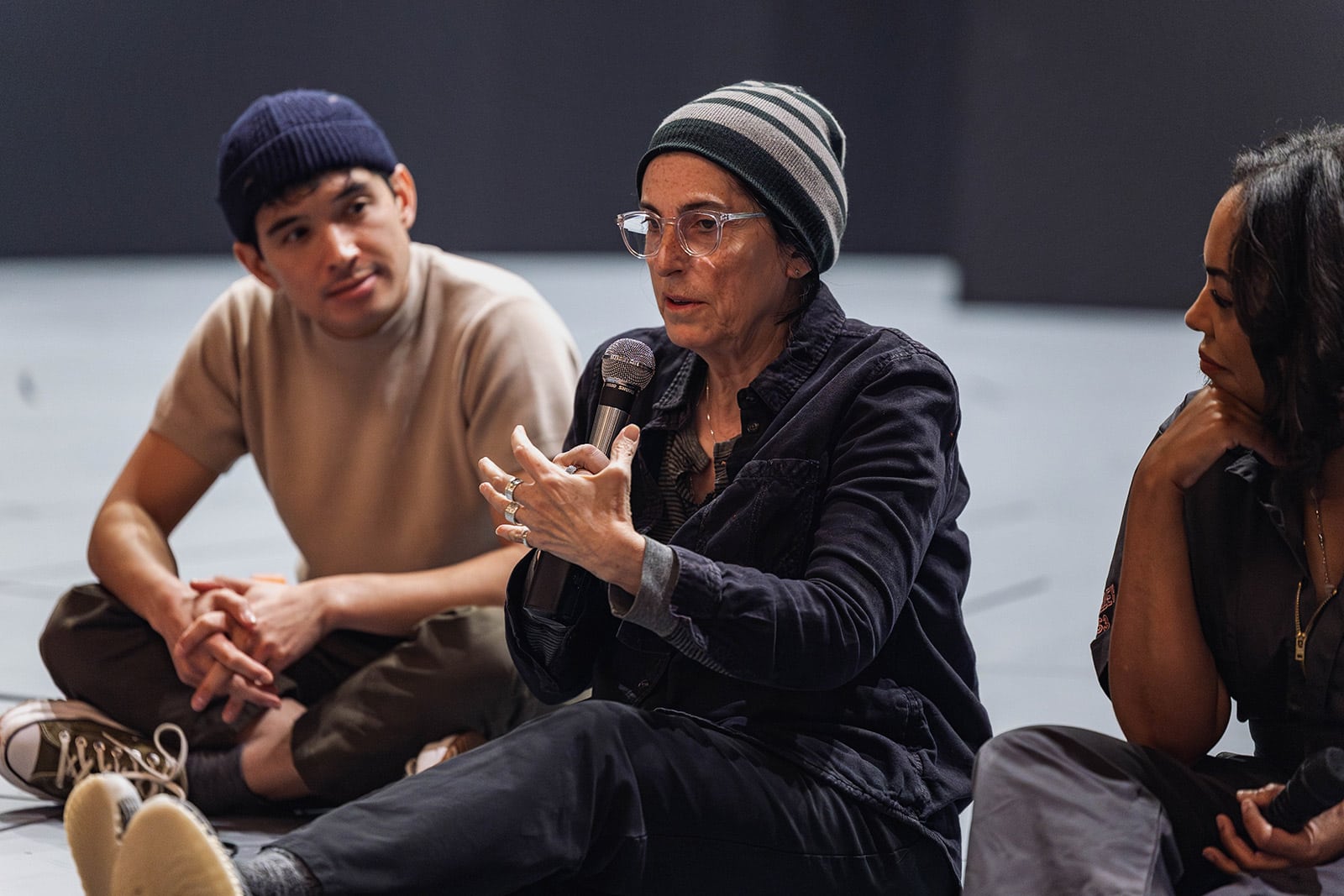 Three cast members sit on the stage, with a woman wearing a beanie speaking into a microphone while two others on each side of her listen