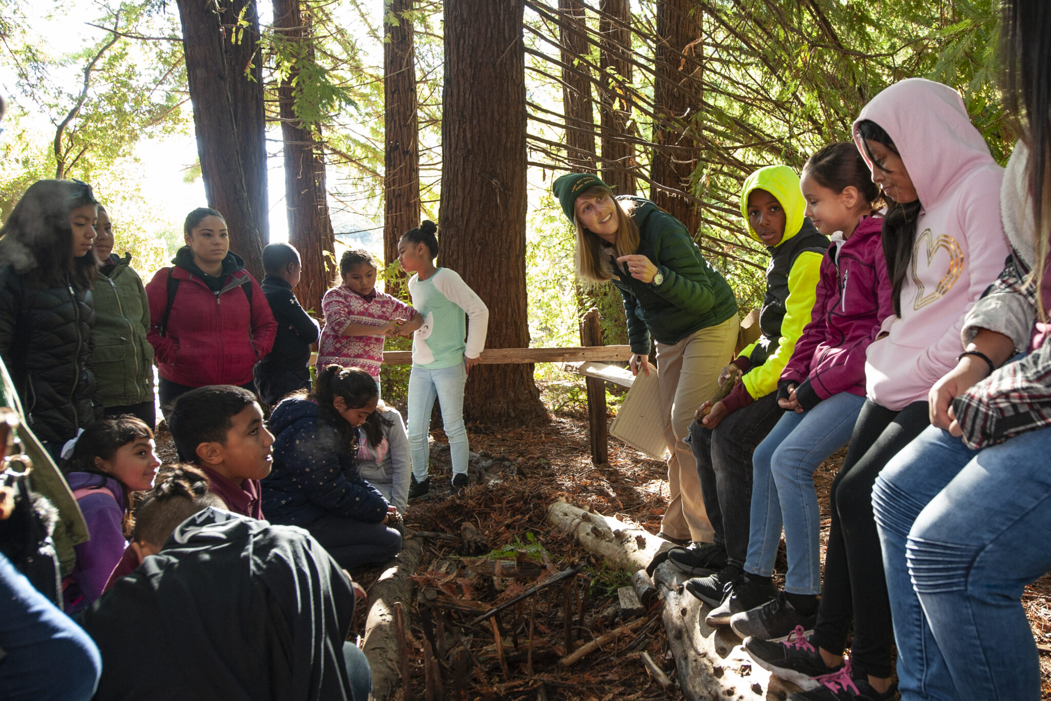 An adult woman with an enthusiastic expression talks to a group of grammar school students sitting on logs in a redwood forest