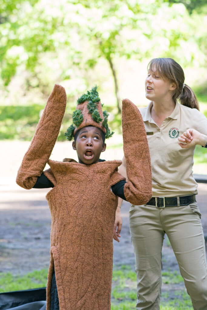 A Black boy wearing a cloth redwood tree costume raises his arms while an adult naturalist in an East Bay Regional Parks uniform points out parts of the tree.
