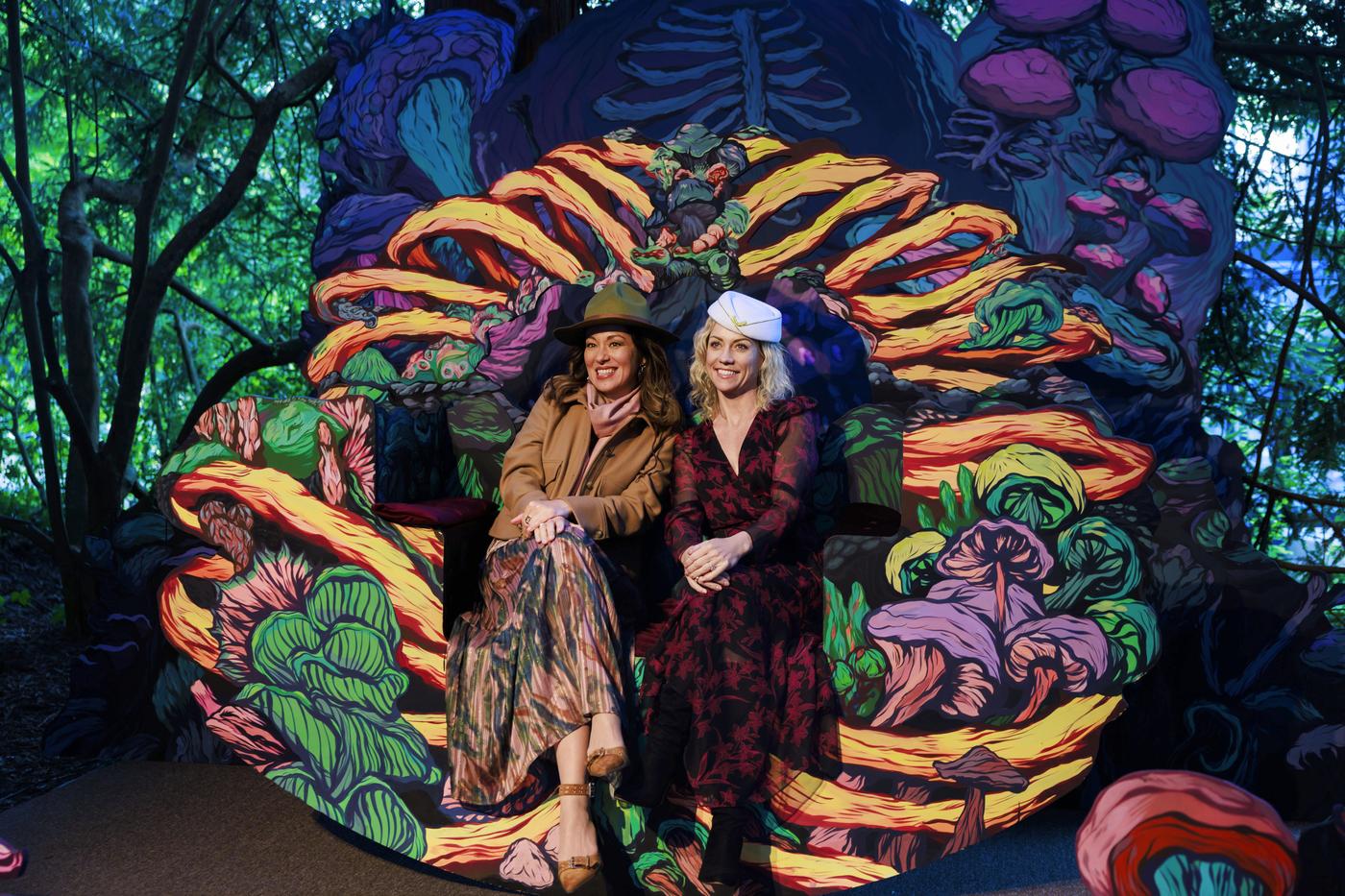 Two women sit on a decorative seat with brightly colored mushrooms and plants