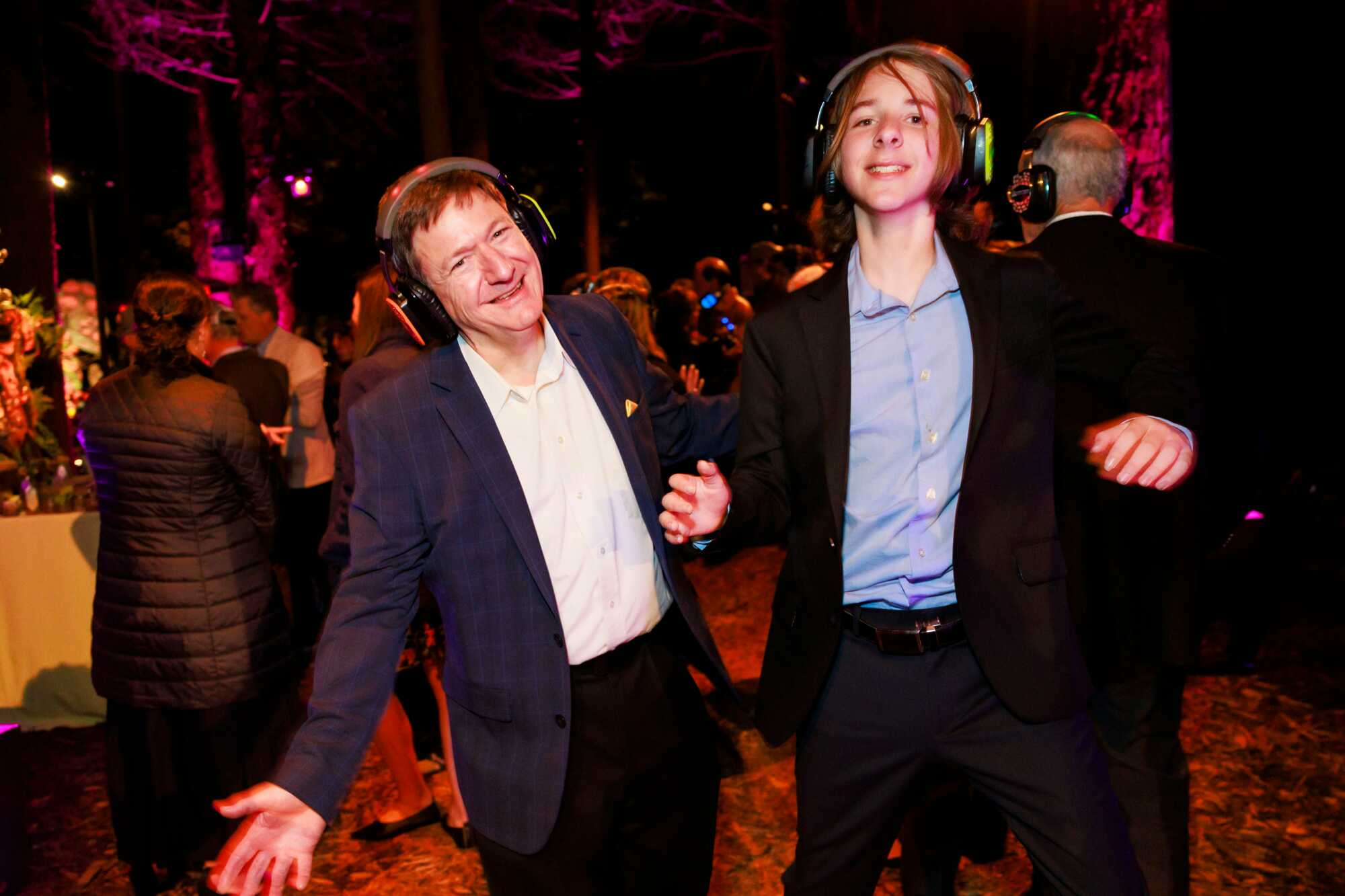 Two men in blazers with over-ear headphones dance against a dark background