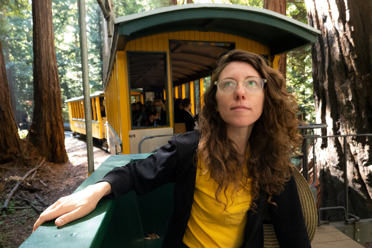 A woman with glasses sits on a locomotive passing through a forest