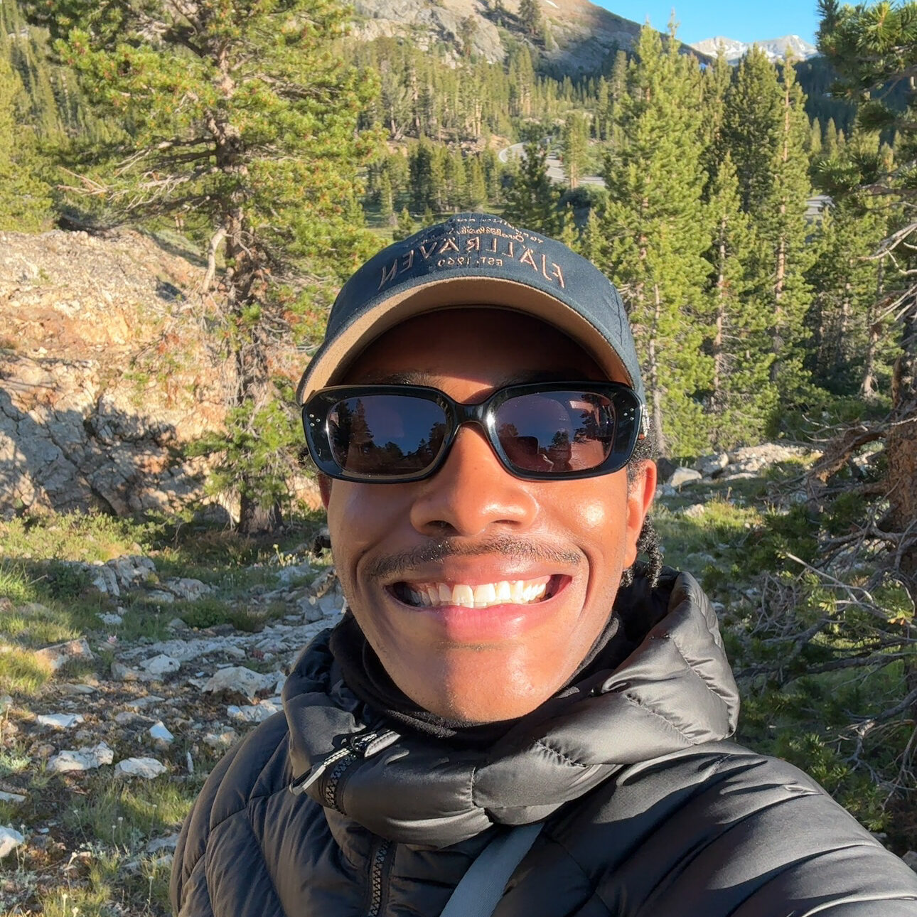 Rahsaan McFarland II, a young black man, wearing sunglasses, a baseball cap and a puffy jacket, smiles into the camera for a selfie while out in the rugged outdoors