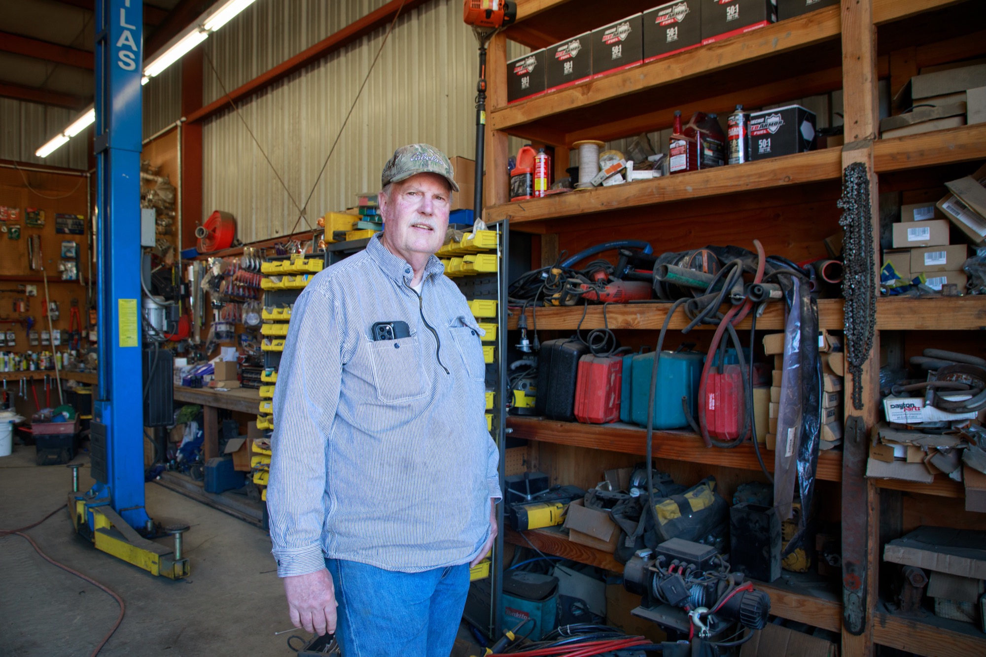 Inside a garage, a man wearing a cap and shirt looks at the camera while surrounded by power tools