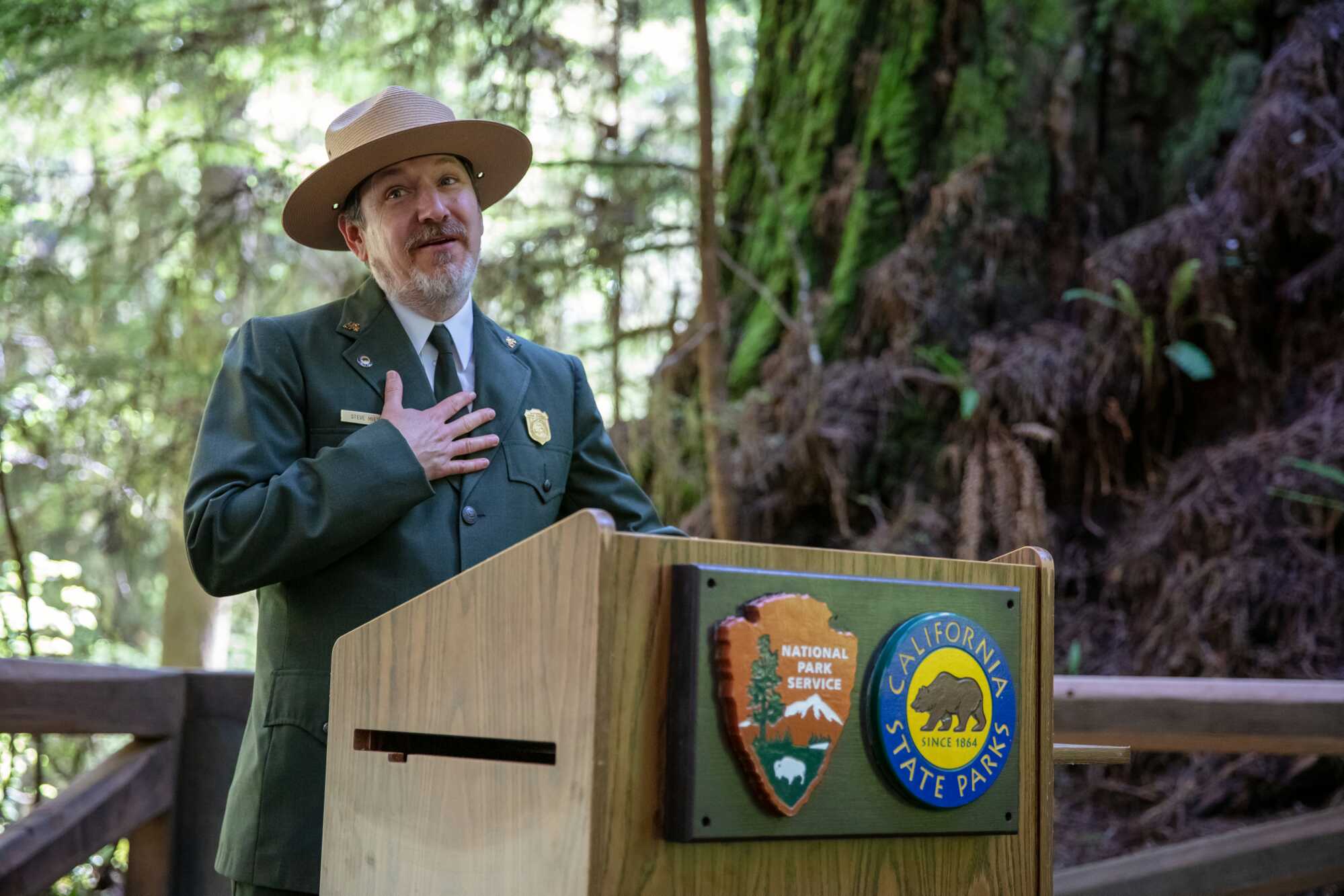 A man wearing a brimmed hat and green blazer stands at a podium in the forest
