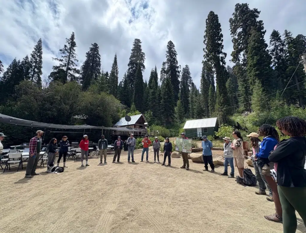 A large group of people stand in a dirt lot preparing for a hike.