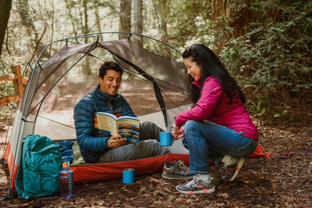 a man shows a book to a woman, both with blue mugs sitting at a tent with a semi-transparent roof in the woods