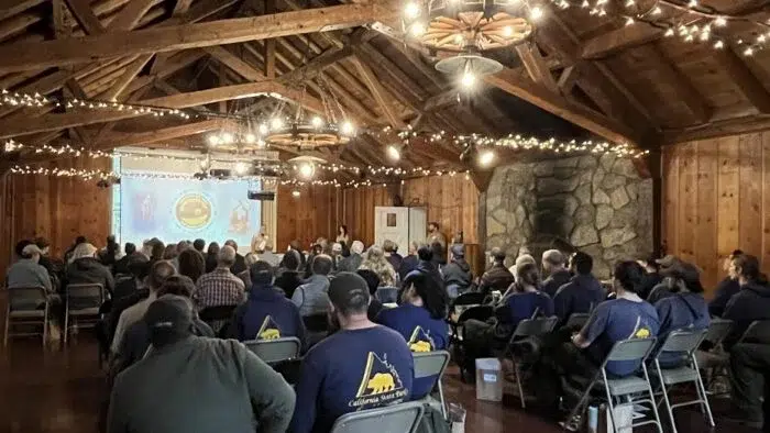 An audience sitting in a high-ceilinged meeting hall faces a large screen and podium at the front of the room.