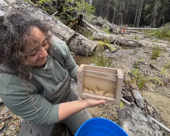 a woman holds a small wooden crate with a glass pane containing small fish swimming in water inside